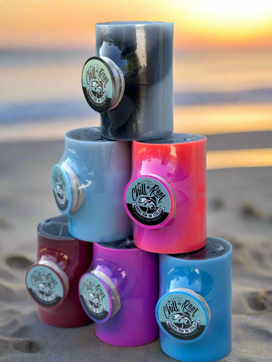 A pyramid stack of six colorful Mystery Color Chill-N-Reel mugs with "Chill-N-Reel®" logos on the beach. The mugs, available in limited edition options, shine in vibrant colors including grey, blue, red, and pink. The background features a blurred sunset over the ocean, creating a serene atmosphere.