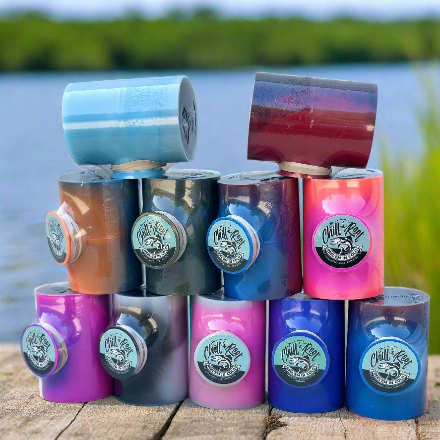 A stack of vibrant Mystery Color Chill-N-Reel wheels by Chill-N-Reel®, including a Limited Edition option, is arranged on a wooden surface with a blurred natural water scenery in the background. The wheels come in various hues—blue, purple, pink, and red—with the brand logo displayed on each wheel.