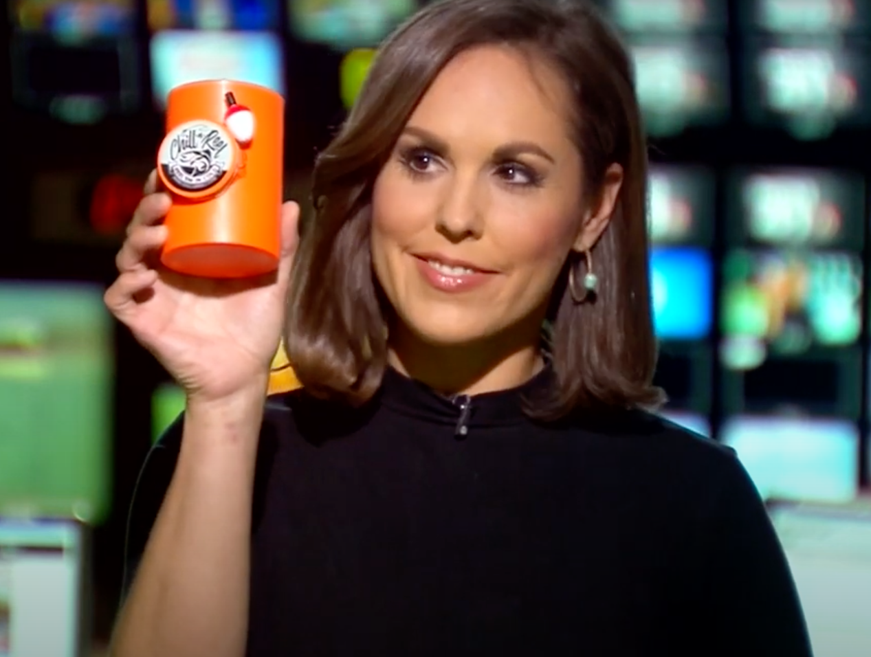 A woman with medium-length brown hair is holding up an orange container with a "Chill-N-Reel" logo on it. She is smiling and wearing a black top. The background is blurred with multiple monitors displaying various images.
