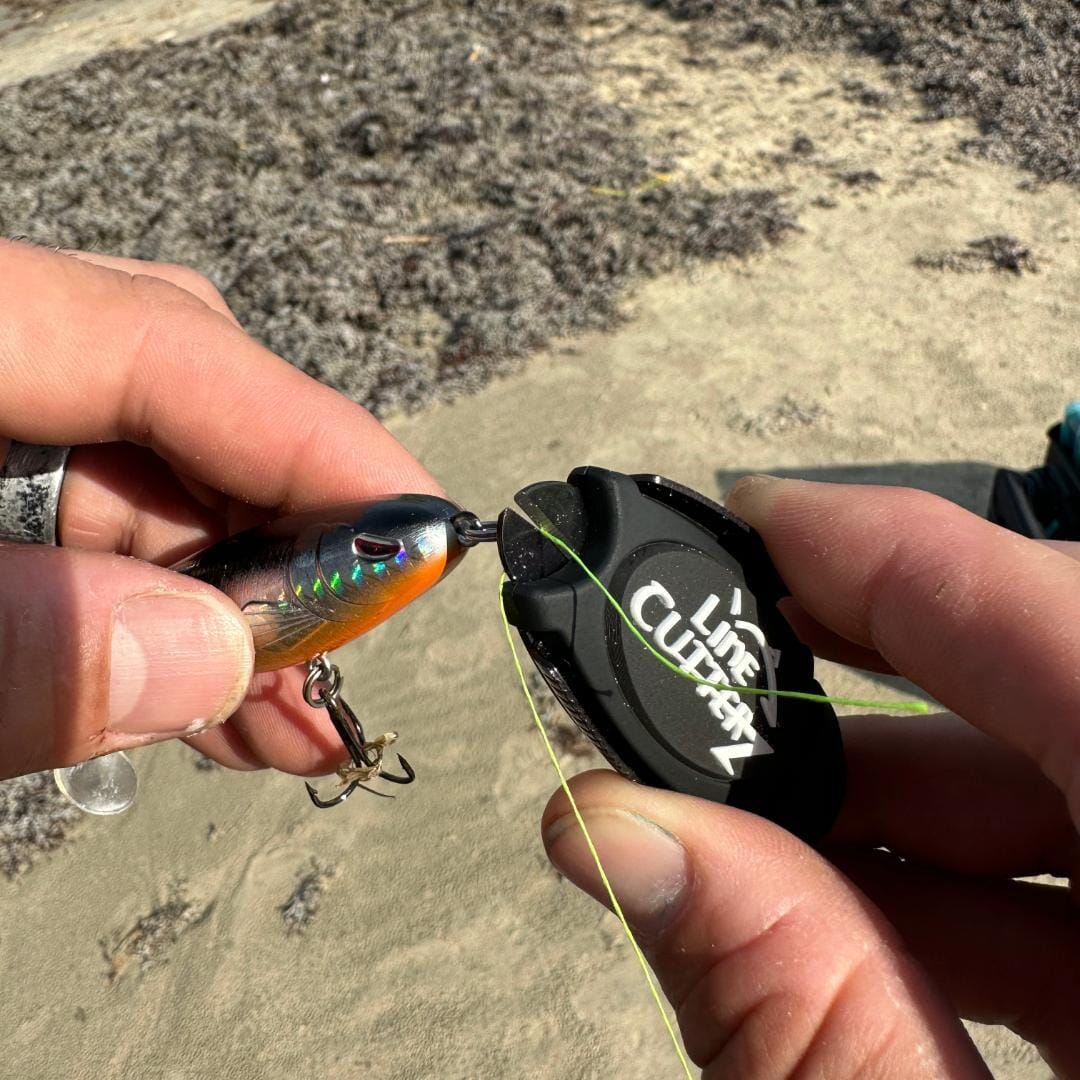 A close-up of two hands cutting fishing line with the Line Cutterz Dual Hybrid Micro Scissors by Line Cutterz, featuring ceramic cutting technology, attached to a lure on a sandy beach background.