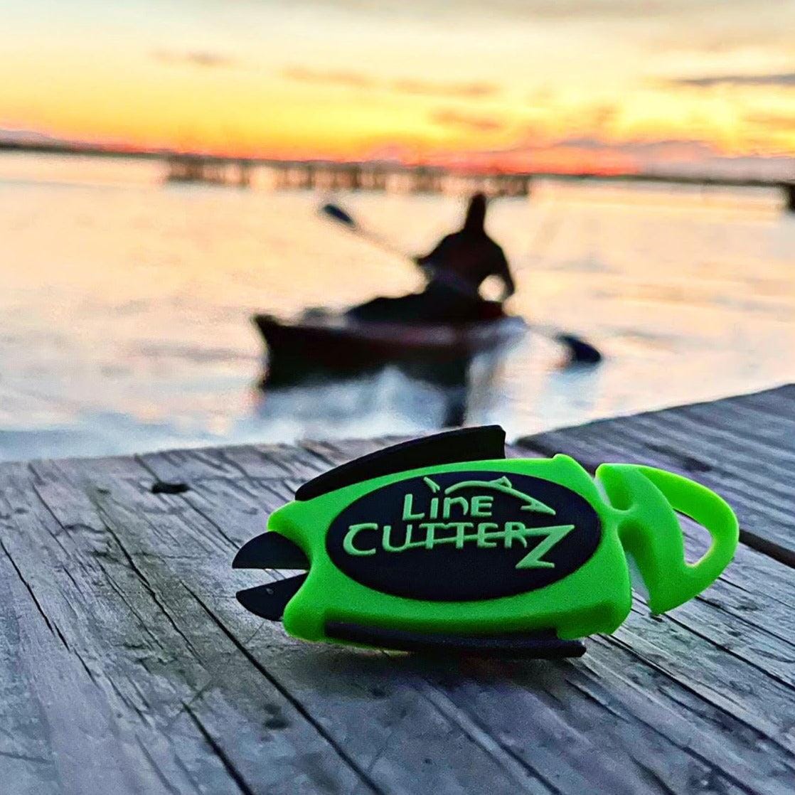 A Line Cutterz Dual Hybrid Micro Scissors by Line Cutterz sits on a wooden surface near the water, while a kayaker is visible in the blurred background at sunset.