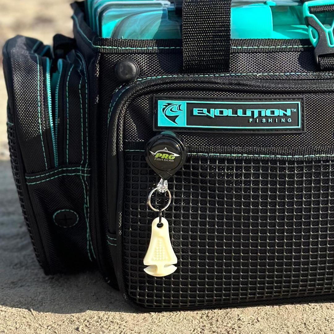 A black Evolution Fishing tackle bag with teal accents sits on sand. A Line Cutterz Ceramic Blade Zipper Pull by Line Cutterz and a white fish hook remover hang from the bag, with clear blue tackle boxes visible inside.