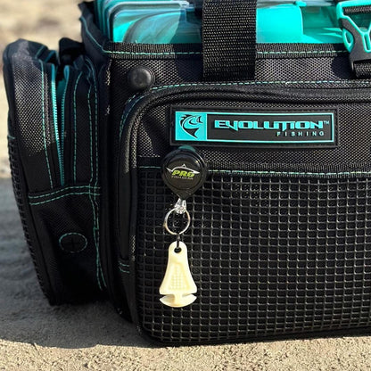 A black Evolution Fishing tackle bag with teal accents sits on sand. A Line Cutterz Ceramic Blade Zipper Pull by Line Cutterz and a white fish hook remover hang from the bag, with clear blue tackle boxes visible inside.