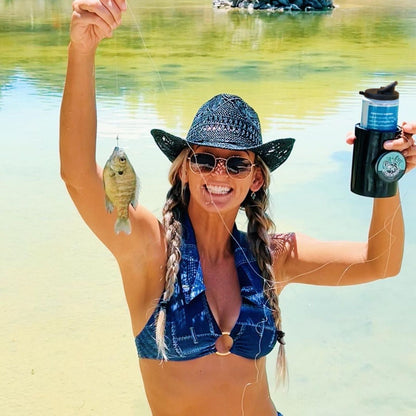 A person with braided hair wearing a blue bikini, sunglasses, and a hat holds up a small fish in one hand and an Original Chill-N-Reel in the other. She is standing in front of a body of water, smiling broadly at the camera, perhaps showcasing her new fun fishing gift—a Chill-N-Reel product.