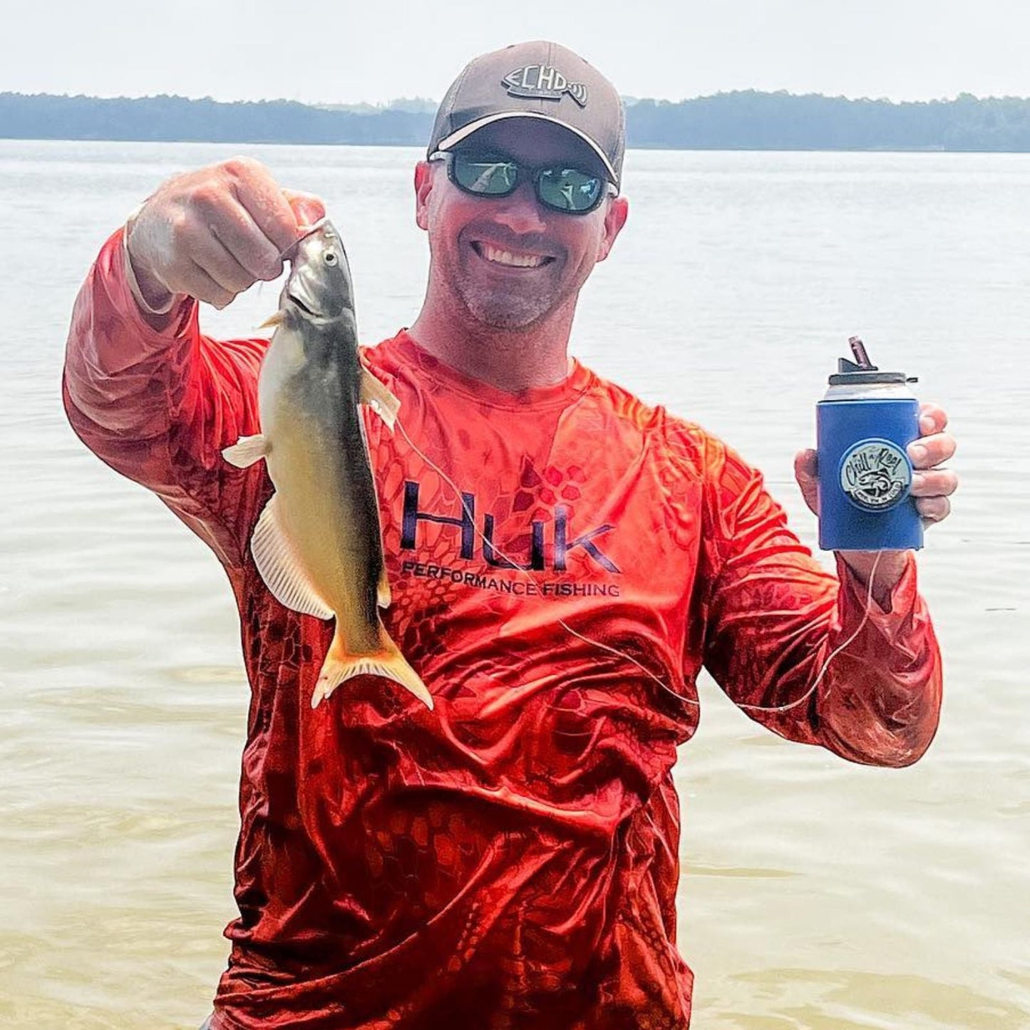 A man in a red fishing shirt and sunglasses holds up a fish he caught while standing in shallow water, smiling with an Original Chill-N-Reel from Chill-N-Reel in his other hand. The background showcases a lake with a tree-lined shore.