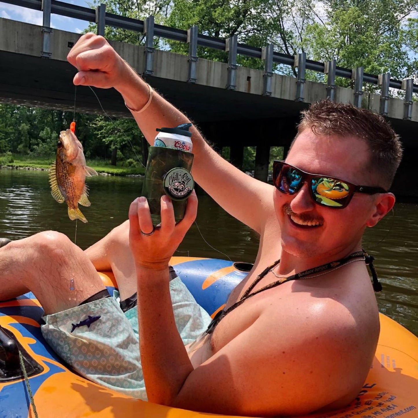 Man on an inflatable tube in a river, smiling, holding a fishing line with a small fish on the hook in one hand and a can of beer wrapped in what looks like a Shark Tank product—an Original Chill-N-Reel by Chill-N-Reel—in the other. He is wearing sunglasses and has short hair, with a bridge and trees in the background.