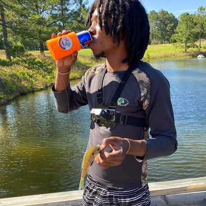 A person with dreadlocks, wearing a dark shirt and striped shorts, holds a small fish in one hand while drinking from an Original Chill-N-Reel by Chill-N-Reel with a blue can in the other. He is standing by a wooden railing near a pond with trees in the background.