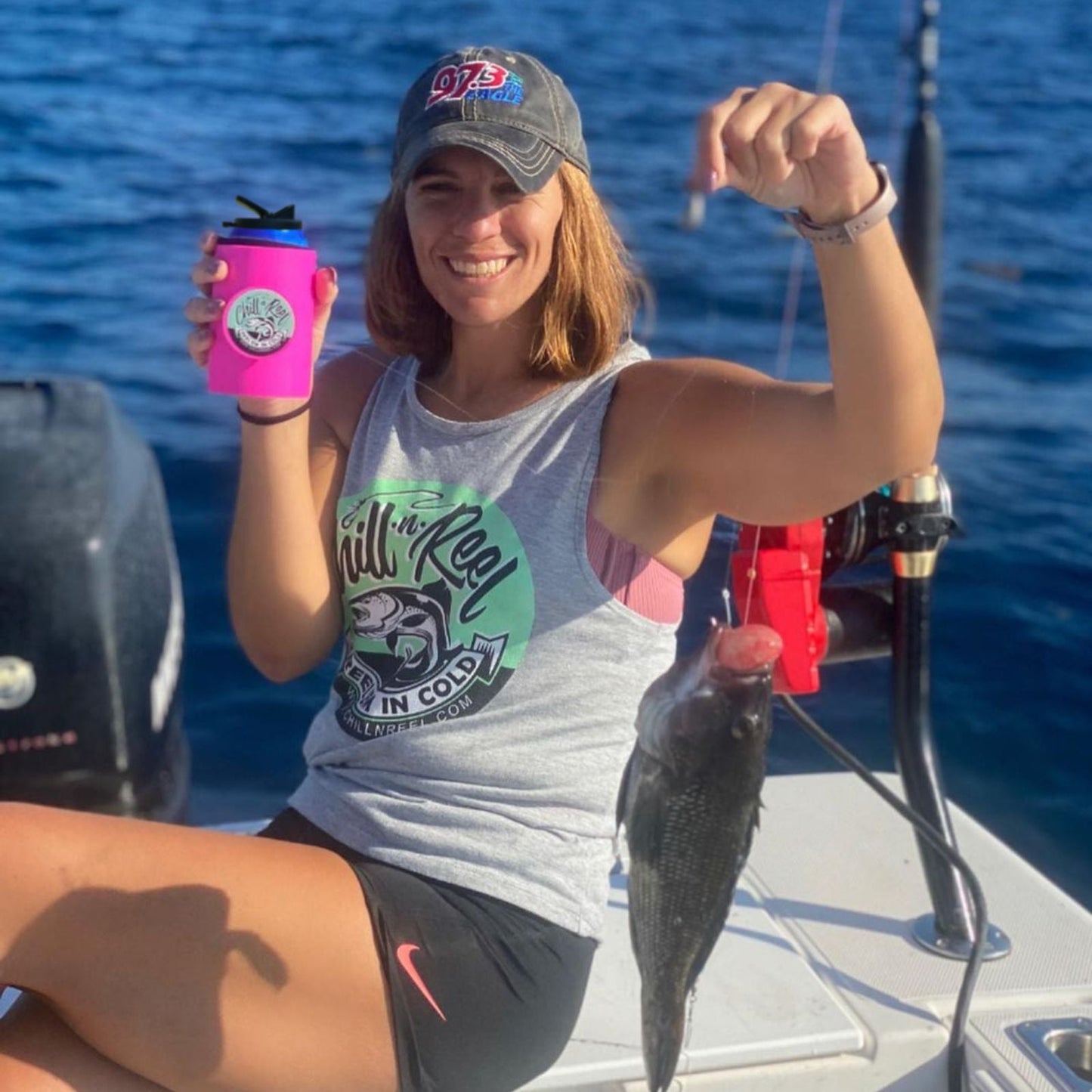 A person is sitting on a boat holding a fishing line with a fish attached in one hand and a fun fishing gift—a Pink Original Chill-N-Reel by Chill-N-Reel—in the other. She is wearing a sleeveless shirt, shorts, and a cap, smiling while enjoying a sunny day on the water.