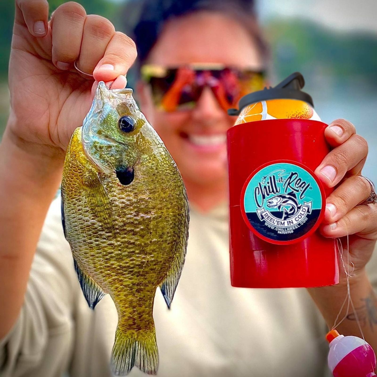 A person holding a small, freshly caught fish in one hand and an insulated red drink holder with a beverage in the other. The drink holder features a fishing-themed design labeled "Chill Reel." The person is smiling and wearing reflective sunglasses.