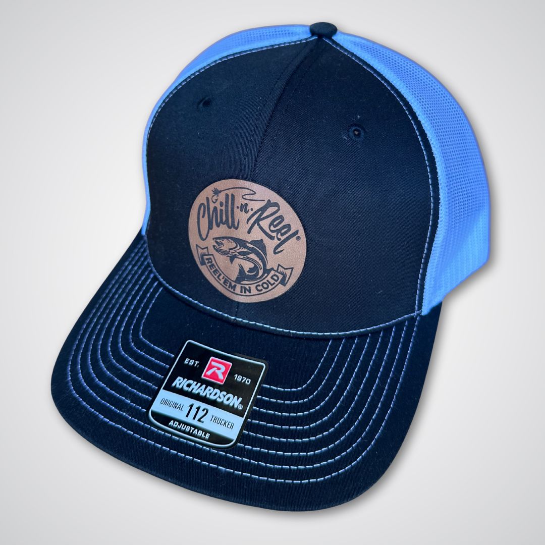 The Chill-N-Reel Trucker Hat by Chill-N-Reel is a black and blue Richardson 112 cap with white stitching, a brown leather patch that reads "Chill n' Reel Reel em in cold" with a fish graphic, and a label on the brim.
