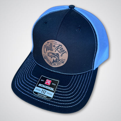 The Chill-N-Reel Trucker Hat by Chill-N-Reel is a black and blue Richardson 112 cap with white stitching, a brown leather patch that reads "Chill n' Reel Reel em in cold" with a fish graphic, and a label on the brim.
