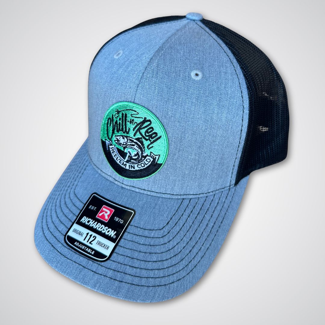 The Chill-N-Reel Trucker Hat features a blue and black design with a mesh back, a green and black fishing patch with fish graphic on the front, and a Richardson 112 sticker label on the brim.