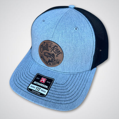 The Chill-N-Reel Trucker Hat by Chill-N-Reel features light gray and black panels, a brown circular front patch with a fish graphic, and a "Richardson 112 Trucker" sticker on the brim.