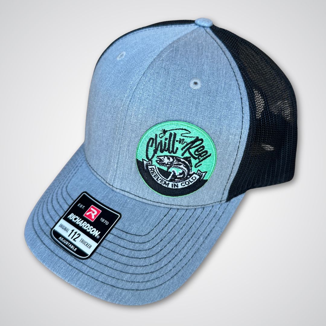 The Chill-N-Reel Trucker Hat by Chill-N-Reel is a gray and black mesh cap with a green circular fish-logo patch on the front and a Richardson 112 tag on the brim, shown against a light gray background.