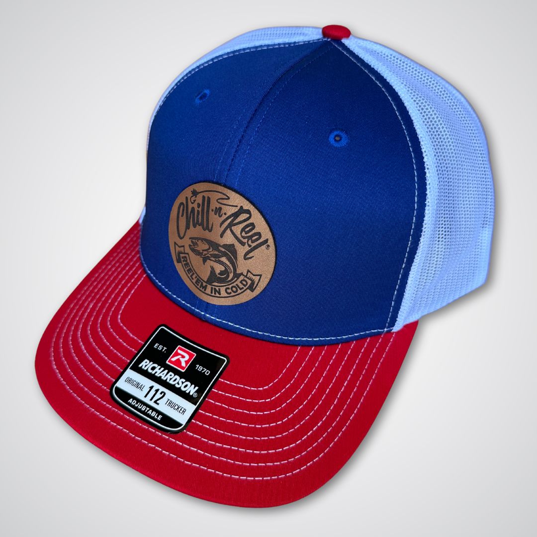 The Chill-N-Reel Trucker Hat features blue and white panels, a red brim, and a round brown Chill-N-Reel patch with a fish on the front. It also has a Richardson 112 sticker for authenticity.