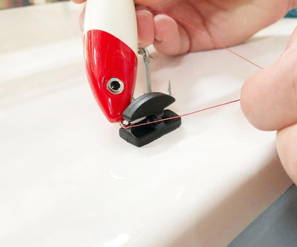 A close-up of a Line Cutterz Ceramic Blade Peel & Stick Flat Mount being used to cut red fishing line threaded through a fishing lure with a red and white head, held by a person's hand.