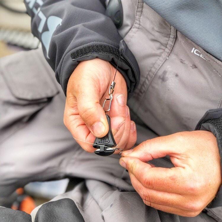 Close-up of hands tying fishing line to a lure, wearing gray waterproof gear with "ICE" on the sleeve and a Line Cutterz Ceramic Blade Zipper Pull by Line Cutterz attached for quick, efficient line trimming.