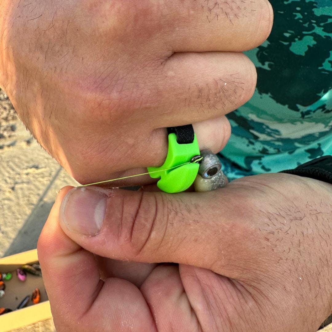 A close-up shows hands threading fishing line through a hook using the Line Cutterz Ceramic Blade Ring by Line Cutterz. The user wears a turquoise patterned shirt, with other fishing accessories visible in the background.