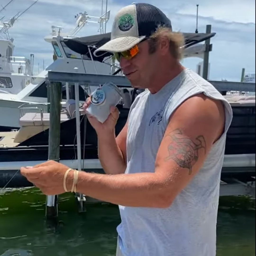 A man with a tattoo on his left arm stands on a dock by the water, holding a drink in his right hand and looking at something in his left hand. He's wearing a sleeveless shirt, a trucker cap, and sunglasses. Several boats are docked in the background.
