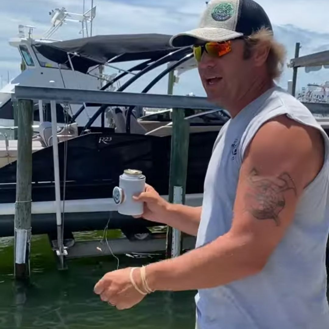 A person wearing a sleeveless shirt with a tattoo on their left arm holds a beverage can wrapped in a holder. They stand near a docked boat, with poles and water visible in the background. The person is wearing sunglasses and a cap.
